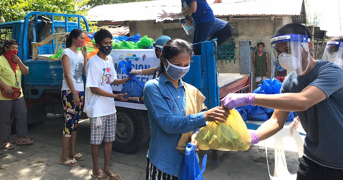 A woman receives a yellow bag of food as a part of UMCOR's Covid-19 response. She, and others around her, are wearing masks. Those delivering food stand in truck beds.
